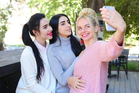 Three wonderful young cute female girl girlfriends make selfie, photo on phone by heart, smiling and laughing in cafe on background trees outdoors. Girls with black hair dressed in white and gray knitted sweaters , girl with blonde hair dressed in pink knの写真素材
