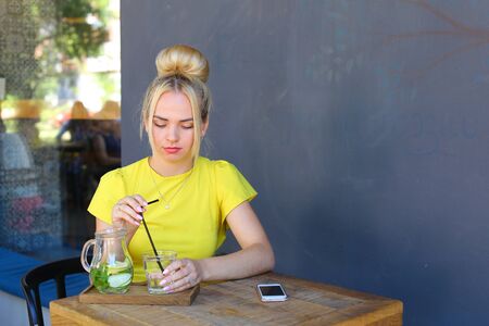 Stunning cute young girl female woman holds glass in hand, pours lemonade and sits at wooden square table against background of gray walls and windows in coffee shop outdoors. Girl with blond hair dressed in short citric dress.Concept of meeting with frieの写真素材