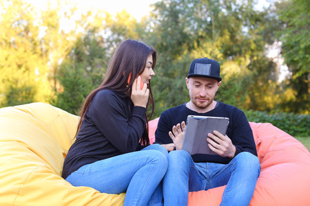 Pair of lovers of young attractive people sitting in soft yellow and red chair, man uses gadget that...