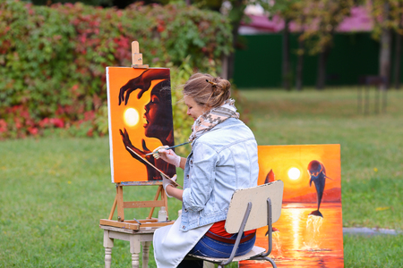 Young cute woman artist  finishes painting which depicts woman who keeps  sun on orange background, sitting with his back to  photographer and  behind wooden easel, holds palette smeared in paint in large park green day outdoors.Girl dressed in red sweateの写真素材