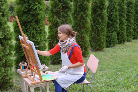 Female artist holds brush and wrote oil on canvas painting, does what likes, interests and hobbies,  preparing picture to order, advertises or art studio for drawing utensils, sits on chair at  wooden easel in large green park outdoors. Girl with brown haの写真素材