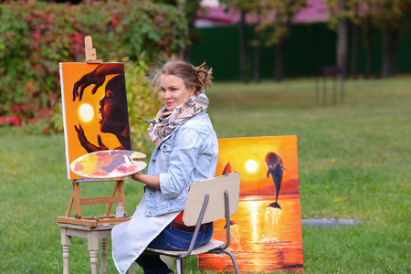 Young cute woman artist  finishes painting which depicts woman who keeps  sun on orange background, sitting with his back to  photographer and  behind wooden easel, holds palette smeared in paint in large park green day outdoors.Girl dressed in red sweateの写真素材