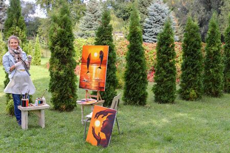 Artist cleans workplace, evaluate work that stands on easel, smiling, posing and looking in camera, standing in full length near paintings in park outdoors on background of coniferous plants in afternoon. Girl dressed in red sweater, dark jeans, white aprの写真素材