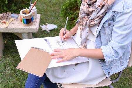 Contemporary Artist Works on Facial Contour of Depicted on Portrait Unknown Girl, Draws Sketch of Fictional Head, Prepares Gift For Friend. Woman Located in Park Outdoors in Background Stands Bench With Colors and Additional Art Materials. Artist Dressed の写真素材