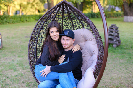 Couple in love swinging on chair in street among  large green park, discussing plans for future, share secrets and jokes, young people hugging each other gently. Guy dressed in black sweater, blue jeans, sneakers and black cap, girl with long black hair dの写真素材