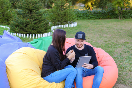 Pair of lovers of young attractive people sitting in soft yellow and red chair, man uses gadget that keeps in arms, female holds plastic card, coupon. Young people cute communicate, plan to travel, choosing interior of apartment, she shows on flyer and guの写真素材