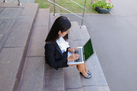 Modern young women, business people, international students holds laptop and study work plan, solve important problems and smiling sits on stairs on background of gray wall of business center outdoors. Woman with black hair, dressed in white blouse and clの写真素材