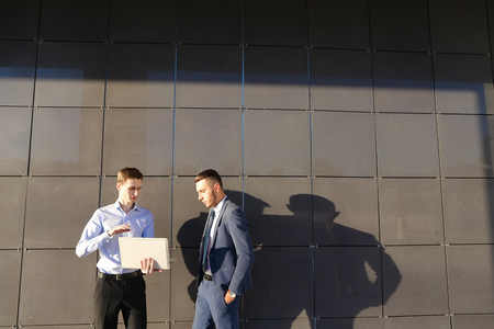 Handsome confident young male man, businessman, student holds laptop in hands and deliver presentations, talks about gadget, advertises new technology to colleague who listening to attentively, and stand on background wall of modern business center outdooの写真素材