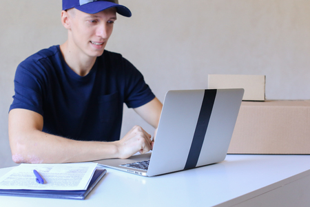 Young Postman Sits at Table Typing at Laptop. Courier Company Accepts Orders Every Day Different Types and Sizes Young Deliveryman European Appearance in Black T-Shirt and Cap Sitting at White Table Typing on Computer and Takes Pen Writing Down Informatioの写真素材
