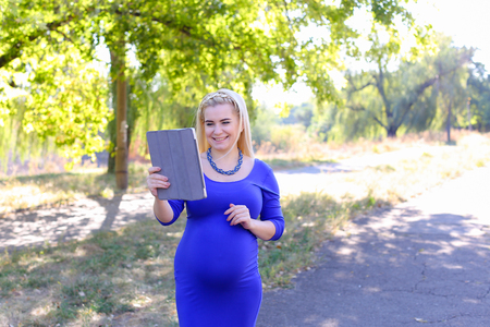 Gorgeous Contemporary Young Feminine Carries Baby Under Heart. Girl Chatters on Skype With Husband About Well-Being or Informs Parents About Anticipated Date of Childbirth. Female With Blond Braided Plait Hair in Front Dressed in Long Blue Dress With Sleeの写真素材