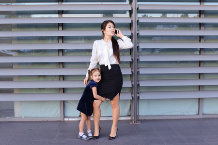 Elegant young female woman, young modern mom, business woman holding mobile phone hand and talking on cellphone, solves problems, negotiates business meeting with partners, spends time girl daughter, talking, smiling at her, little girl hugging her motherの写真素材