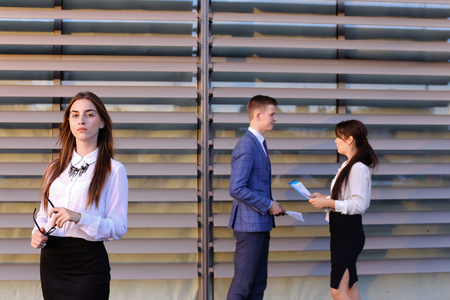 Successful young adult  business lady, student rests, took break from work, watching, smiling and posing at camera and stands on background of colleagues who discuss workflows, consider paper near of business center outside. Girls with light brown and darの写真素材