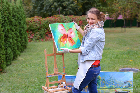 Female European appearance artist evaluates picture and removes from easel, holds and posing with picture which depicts colorful butterfly, smiling and standing in full growth on background of lawn and angered by conifers in large park in open air. Girl dの写真素材