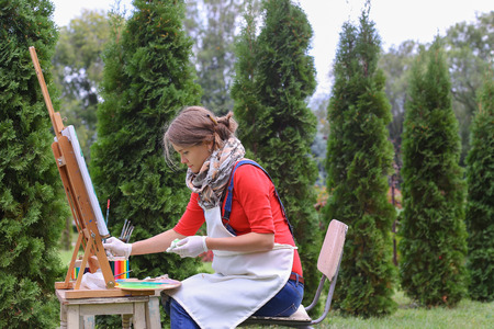Female artist holds brush and wrote oil on canvas painting, does what likes, interests and hobbies,  preparing picture to order, advertises or art studio for drawing utensils, sits on chair at  wooden easel in large green park outdoors. Girl with brown haの写真素材
