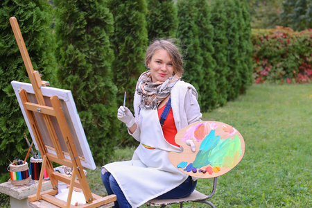 Cute female painter posing, laughing and in front of camera , draws and smiling with brushes and colorful palette in hand, sits on chair near easel in green park outdoor.  Girl with brown hair collected in beam dressed in light jacket, red jacket, dark deの写真素材