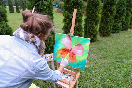 Young girl European appearance looks at drawing of Multicolored butterflies, which painted with oil paints and estimates finalizing remaining details. Stands near wooden easel with brush in hands on background art materials and coniferous trees in park ouの写真素材