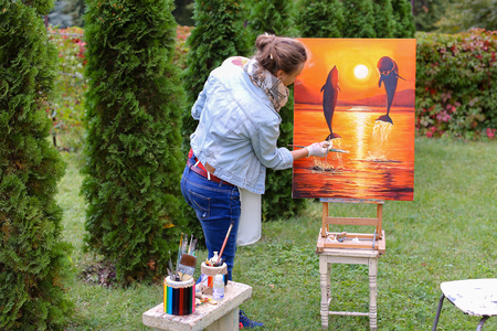 Pretty attractive artist finishes painting which depicts dolphins jumping out of  water on orange background, stands with back to photographer and behind wooden easel, holds palette smeared in paint in large green park day outdoors.Girl dressed in red sweの写真素材