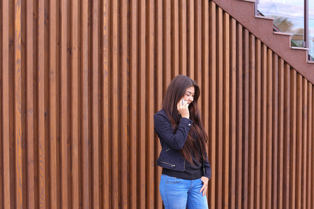 Cute girl with long hair cute talking on phone with friend or loved one, make an appointment. Women dressed in black sweater, blue jeans, black boots on platform and top dressed in jacket standing on background of brown wood panel walls near restaurant ouの写真素材