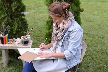 Contemporary Artist Works on Facial Contour of Depicted on Portrait Unknown Girl, Draws Sketch of Fictional Head, Prepares Gift For Friend. Woman Located in Park Outdoors in Background Stands Bench With Colors and Additional Art Materials. Artist Dressed の写真素材