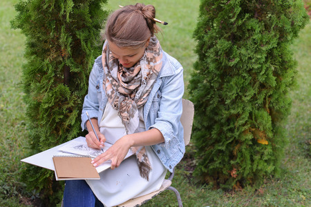 Contemporary Artist Works on Facial Contour of Depicted on Portrait Unknown Girl, Draws Sketch of Fictional Head, Prepares Gift For Friend. Woman Located in Park Outdoors in Background Stands Bench With Colors and Additional Art Materials. Artist Dressed の写真素材