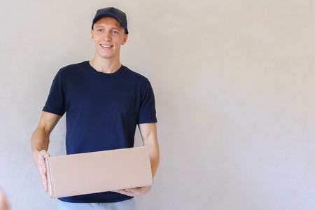 Deliveryman Standing in Room, Glue Tape Box and Takes Parcel in Hands and Smiling . Postman European Appearance in a Black T-Shirt and Cap Stands Near the Desk in the Office With White Walls and Unassisted Packaging Boxes, Adhesive Glue Tape on a Cardboarの写真素材