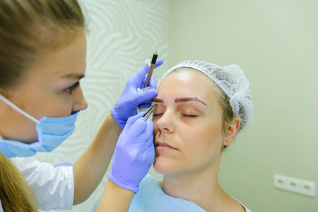 Successful female browmaker consult client girl completes registration and drawing eyebrow hairs and antiseptic wipes. Master dressed in white robe, blue rubber gloves and face protection gauze bandage. Concept of mikrobleyding eyebrows micropigmentation の写真素材