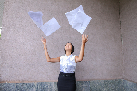 Pretty asian girl smiling and posing, holds paper in hands and looking away on background of wall of buildings outdoors. Woman with black hair, dressed in white blouse and classic skirt. Concept of business meeting, business communication, successful peopの写真素材