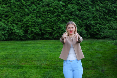 Slender blonde curves and shows gesture "Bad" grieved and shows evaluation of product, does not recommend fitness club or restaurant. Women with long blonde hair dressed in bright blouse, beige jacket and blue pants, standing on background of green grass の写真素材