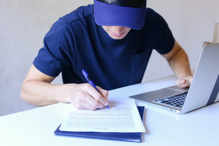 Mailman Sitting in Office on Table and Typing at Laptop, Taking Pen and Writes Information on Document. Young Student Works as Courier and Successfully Performs Work, Worker Has European Appearance in Black T-Shirt and Cap Sitting at White Table Typing onの写真素材