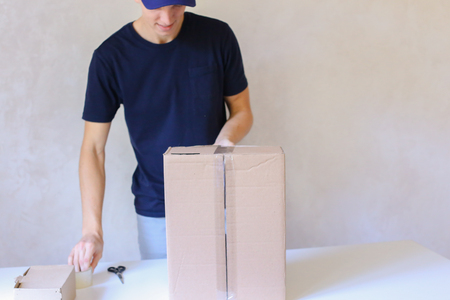 Deliveryman Standing in Room, Glue Tape Box and Takes Parcel in Hands and Smiling . Postman European Appearance in a Black T-Shirt and Cap Stands Near the Desk in the Office With White Walls and Unassisted Packaging Boxes, Adhesive Glue Tape on a Cardboarの写真素材