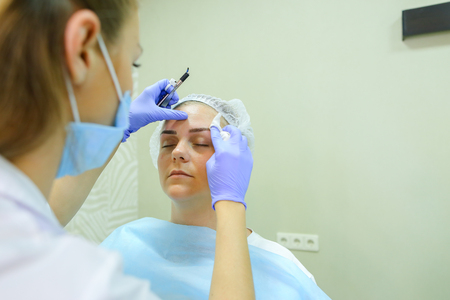 Successful female browmaker consult client girl completes registration and drawing eyebrow hairs and antiseptic wipes. Master dressed in white robe, blue rubber gloves and face protection gauze bandage. Concept of mikrobleyding eyebrows micropigmentation の写真素材
