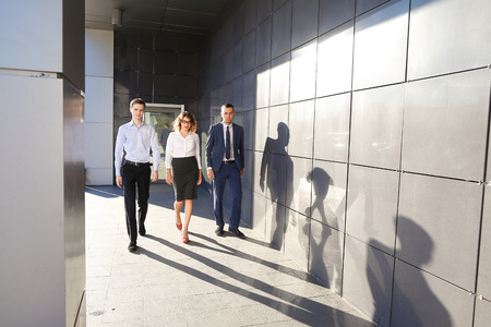 Beautiful successful energetic people, two guys and girl, young businessmen, students confident step forward, smiling and looking at camera on background of gray wall outdoors business center. One of guys dressed in blue classic suit, white shirt with tieの写真素材