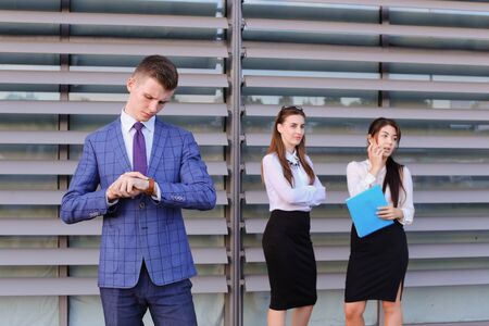 Handsome confident young male guy businessman, office worker, student posing in front camera, looking at  watch, late for business meeting, posing for shoot of modern mens business clothing catalog and stands  background of colleagues, two attractive younの写真素材