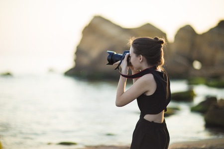 Young woman photographer holding camera and looking into frame. Girl dressed in black jumpsuit, European appearance. Concept of feminine beauty,  work photographer tourist with camera on vacation or trip.の写真素材