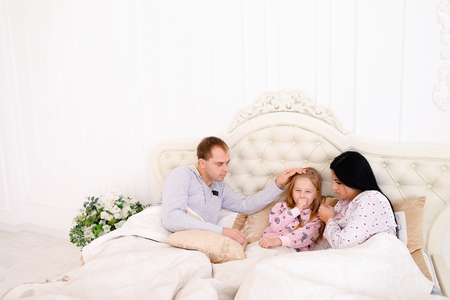 Women caring sick child, wiping nose with tissue. Child in bed with mom and dad. Young couple caring of their daughter. Woman with long black hair, brown man and daughter blonde European appearance against white interior. Concept of family life, common coの写真素材