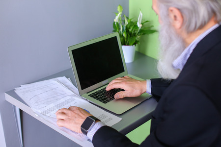 Portrait of male retiree who prints notebook on keyboard and sings report on company's work, looks...