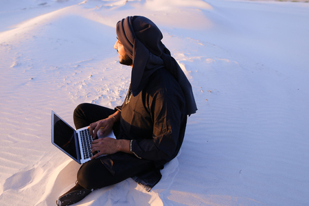 Joyous male Arab sits on sand at computer. young man engaged in scientific work or writes article typing on keyboard of gadget, relaxes sitting in silence of bottomless desert against blue sky on summer evening. Swarthy Muslim with short dark hair and beaの写真素材