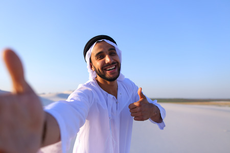 Close-up portrait of attractive Muslim guy holding camera with hand, aiming at himself and smiling with broad smile. Young sheikh guy shows gestures with thumb up and everything okay, standing among bottomless desert with hills of white sand on clear evenの写真素材