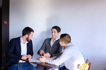Three young men who are held discuss important questions about work and general business. Guy with curly hair talking on phone and hands receiver to seated man, who then conducts dialogue on mobile, sitting in gray modern office at table. Guy with dark cuの写真素材