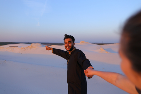 Young Muslim man leads sweet girl by hand, chatting with young woman and smiling, which goes for male. Lovers walk through expanses of sandy desert at sunset under open blue sky. Woman with fair hair tied in tail dressed in light t-shirt and blue jeans, dの写真素材