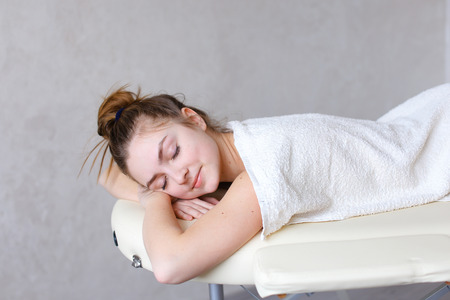 Pretty European woman looks relaxed after receiving procedures with smile on face, turns and poses on camera, lying on white couch, covered with beige terry towel in beauty salon. Concept of relaxation and recreation, variety of restorative massages, cosmの写真素材