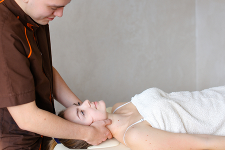 Experienced doctor professionally kneads neck and conducts therapeutic procedure for neck, removes discomfort of young woman who lies on couch, covered with terry towel. Man of European appearance with short haircut dressed in brown medical uniform from sの写真素材