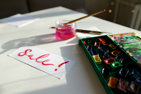 Bright inscription for sale sale, written in pink watercolor paints and brush, which lies on white table in art studio. Concept of fine arts and bright colors, beautiful writing, calligraphy and lettering, materials and tools, craftsmanship and hobbies, sの写真素材