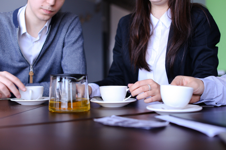 Young wealthy managers of firm, two men and young woman communicate at expense of work and joke professional jokes, girl pours green tea in mugs and workers discuss past working day, sitting in evening cafe. European-looking guy with fair hair and short hの写真素材