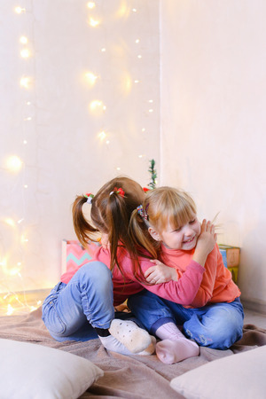 Funny little girls smile and pose in camera, laugh and communicate, hug each other and sit on rug in bright room with New Years small Christmas tree and garland on wall. Blond girl of European appearance with tails on sides dressed in orange sweatshirt wiの写真素材