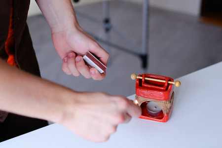 Male therapist doctor holds matches in his hands and lights aromatic candle in special stand, on which lies wooden stick and pours oil with pleasant relaxing odor. Guy dressed in brown medical uniform and in cosmetology room. Concept of profession of massの写真素材