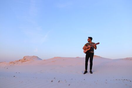 Young Muslim male performer and singer walks through sandy desert with guitar in hands, stops and begins to play on spot on musical instrument, rejoices and smiles, sings songs about love on warm summer evening at sunset. Swarthy man with dark hair and shの写真素材