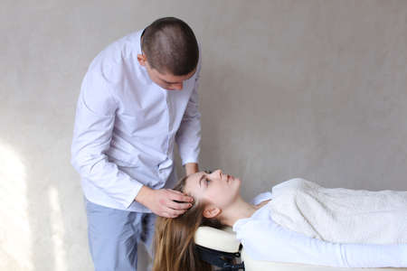 Educated cosmetologist conducts procedure to relieve tension in head and gives advice on care for womans pretty woman who lies on beige couch in medical office. Man of European appearance with short haircut dressed in light gray medical uniform from shirtの写真素材