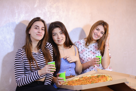 Three wonderful women having fun, ordered large fragrant pizza and try, laughing and talking on girlish themes, discussing guys sitting on floor in bright bedroom at night. women with smile posing and lighting spark of Bengal. European-looking girl with rの写真素材