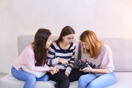 Attractive girls hold modern devices, VR glasses and inspect, study possibilities of gadget and communicate with each other, sitting on gray sofa in room during day. European-looking girl with medium-length blond hair dressed in pink blouse and jeans, secの写真素材